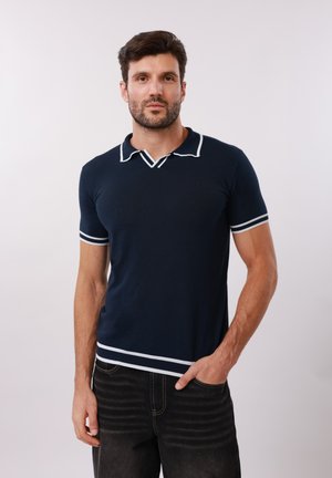 Homme aux cheveux courts et foncés avec une barbe, portant un polo bleu marine avec une bordure blanche et un jean noir, main dans la poche, fond neutre.