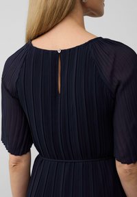 Robe plissée navy à manches courtes et encolure ronde au dos, avec une ouverture en forme de trou de serrure fermée par un petit bouton.