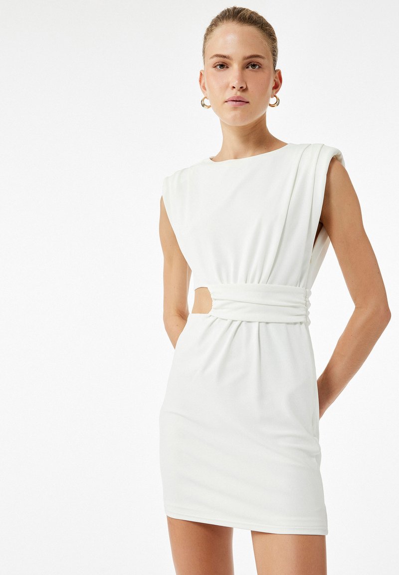 Hvid kjole lavet af blød stof, med bådudskæring, draperede ærmer, en indsnævret talje med udskæringsdetalje og en tætsiddende silhuet.