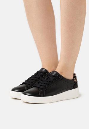 Sorte lædersneakers med tykke hvide såler og leopardmønstret hældetalje båret på bare fødder mod en hvid baggrund.