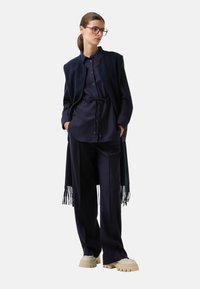 Vrouw in een marineblauwe shirt en broek met een bijpassend mouwloos lang vest en beige chunky platform schoenen, staand met de handen in de zakken.