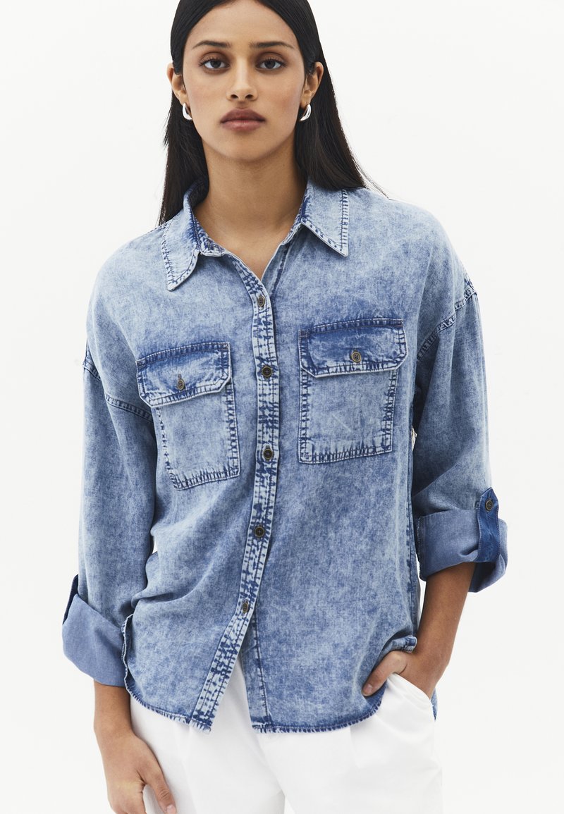 Ung kvinde iført en falmet blå denim skjorte med op-rullede ærmer og hvide bukser, stående med den ene hånd i lommen.