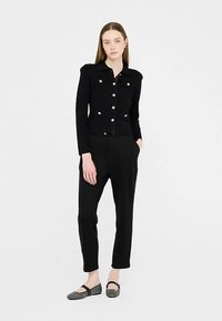 Juoda aptempianti striukė su aukso spalvos sagomis, ilgomis rankovėmis ir struktūruota apykakle, derinama su juodomis kelnėmis ir raštuotomis lakuotomis avalynėmis.