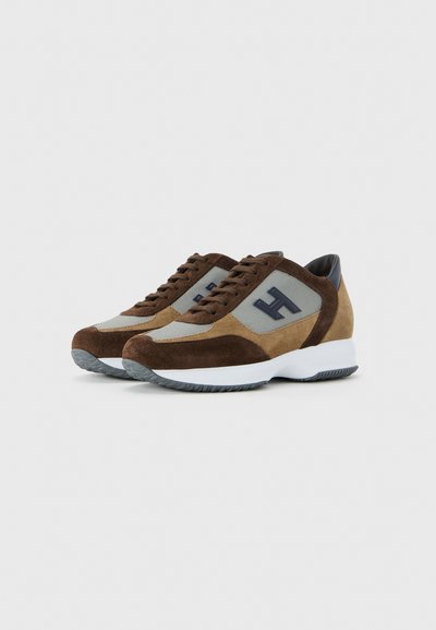 Pair of brown and beige suede sneakers with mesh panels, lace-up detail, dark blue “H” logo on sides, and white rubber soles.