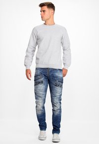 Sweatshirt gris à col rond, porté avec un jean bleu déchiré présentant des poches zippées et des accents noirs, complété par des baskets blanches.