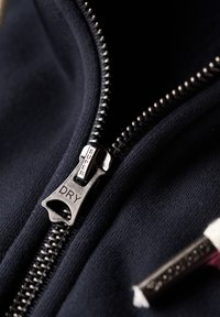 Felpa blu con zip, dotata di un tirazip in metallo etichettato "SUPER" e "DRY", che mostra un tessuto testurizzato e cuciture visibili lungo i bordi.