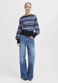 Stickad tröja med ett zigzag-mönster i blått, grått och svart; har långa ärmar och en ribbad svart midja. Matchas med vida jeans.