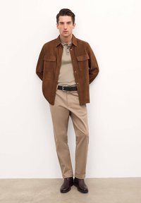 Chaqueta de ante marrón sobre una polo beige, combinada con pantalones beige y botas marrones oscuras. Presenta dos bolsillos en el pecho y un cinturón negro.