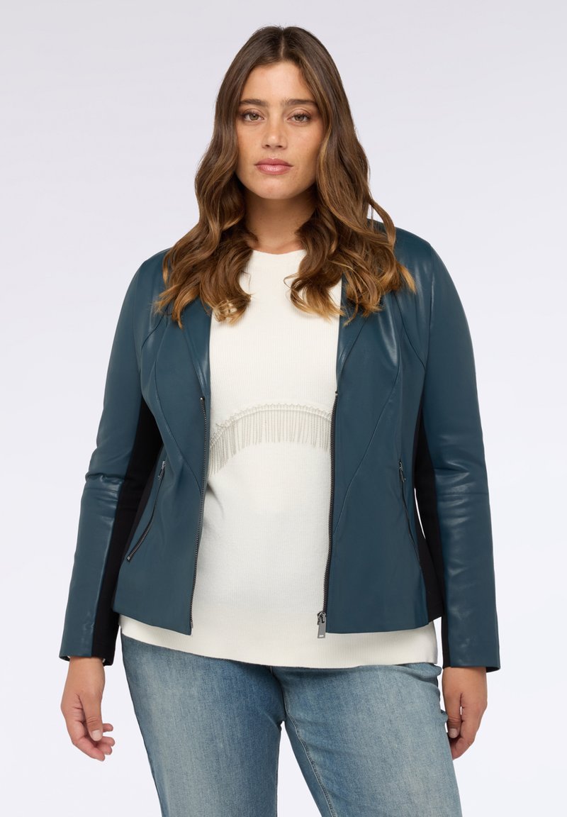 Tyrkysová koženka s přiléhavým střihém, zipem vpředu a černými bočními panely, kombinovaná se světlým topem a modrými džínami.