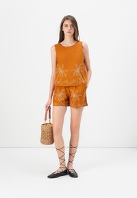 Oranges ärmelloses Oberteil und Shorts-Set mit weißer Palmenstickerei. Das Model hält eine geflochtene Tasche und trägt schwarze Schnürflats.