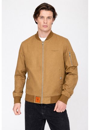 Mann trägt eine beige Bomberjacke mit Reißverschluss, einem Brustemblem, einer Reißverschlusstasche am Ärmel und einem orangefarbenen Etikett am Saum, steht vor weißem Hintergrund.