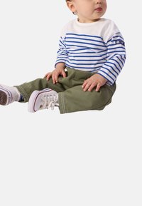 Λευκό πουκάμισο με μακρύ μανίκι και οριζόντιες μπλε ρίγες, συνδυασμένο με παντελόνι ελιάς. Αθλητικά παπούτσια με λευκή βάση και κόκκινα στοιχεία.