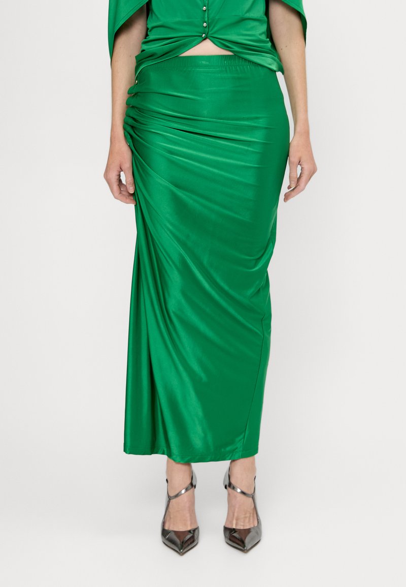 Grøn satin maxi-nederdel med draperet detaljering, elastisk talje og glat tekstur. Sølv høje hæle komplementerer outfittet.
