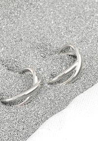 Silvermetallörhängen med en böjd, öppen design. Slät textur med eleganta, polerade ytor och spetsiga ändar. Omgivna av en glittrande bakgrund.