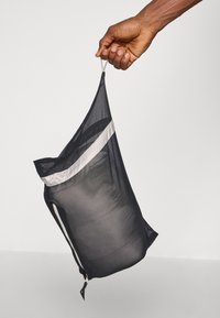Sac din plasă negru cu șnur, având o dungi albă, ținut în mână. Prezintă un fermoar și o textură semi-transparentă.