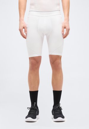 Muž v bielych kompresných šortkách, čiernych ponožkách a čiernych športových topánkach stojaci pred obyčajným svetlým pozadím.