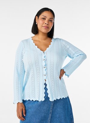 Femme aux cheveux châtains mi-longs porte un cardigan bleu clair à bord festonné avec des boutons en perles et une jupe en jean bleue, posant avec la main sur la hanche.