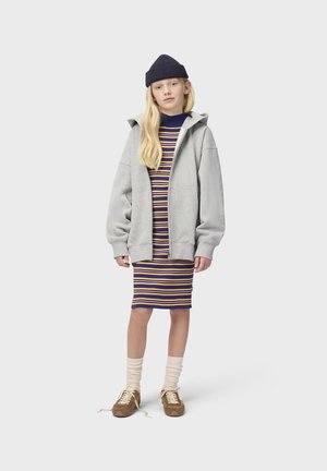 Dziewczyna w granatowej czapce typu beanie, szarej bluzie rozpinanej na suwak, pasiastych sukience do kolan, kremowych skarpetkach oraz brązowych trampkach z rozwiązanymi sznurowadłami na jednolitym tle.
