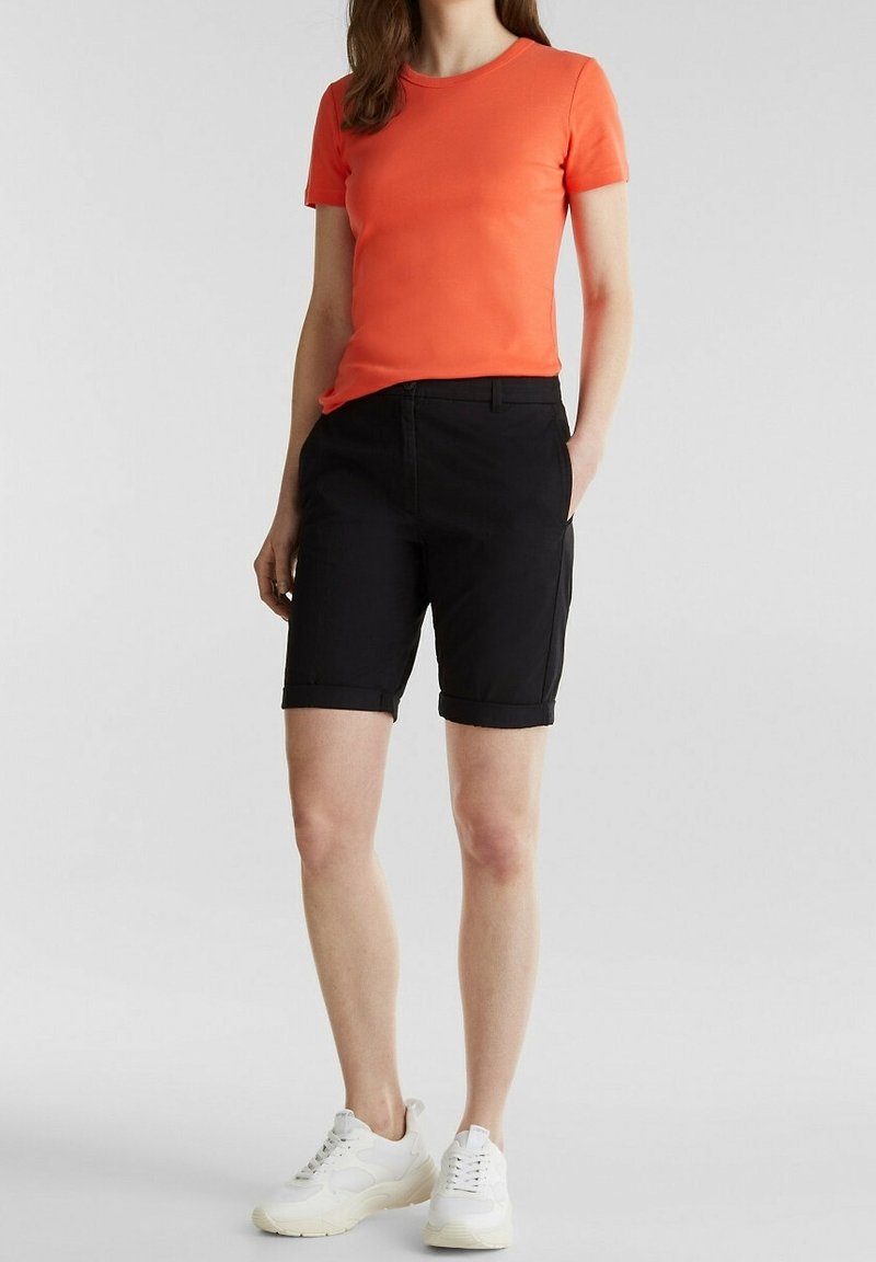 Femme portant un t-shirt orange vif à manches courtes, un short noir jusqu’aux genoux et des baskets blanches, debout avec une main dans la poche.