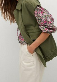 Olive green bezrękawnik na koszulę w kwiaty z różowymi i białymi motywami. Spodnie w kolorze kremowym, z teksturowanej tkaniny.
