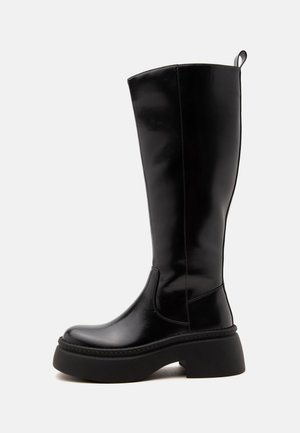 Botte noire haute en cuir avec semelle épaisse et talon bloc, profil latéral sur fond blanc, présentant une languette de tirage en haut à l'arrière.
