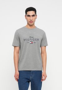 Mladic nosi sivo Tommy Hilfiger majicu s logotipom i tekstom 1985, uparen s plavim trapericama, stoji naslonjen na bijelu pozadinu.