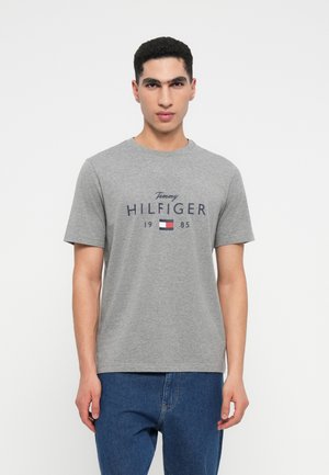 Tânăr îmbrăcat cu un tricou gri Tommy Hilfiger cu logo și textul 1985, asortat cu blugi albaștri, stând pe un fundal alb.
