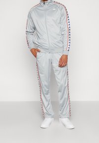 Homme portant un survêtement gris clair avec des bandes aux logos bleu et rouge sur les manches et le pantalon, associé à des baskets blanches.