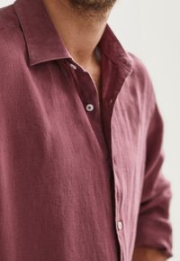 Burgundowa lniana koszula z kołnierzykiem na guziki, widocznymi guzikami i miękkim, teksturowanym materiałem. Wydaje się, że ma luźny krój z podwiniętymi rękawami.