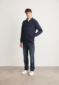 Felpa con zip navy, dotata di cappuccio regolabile, tasca a marsupio e dettagli con logo, abbinata a jeans scuri e sneakers bianche su uno sfondo neutro.