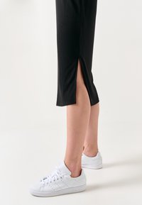 Jambes portant un pantalon noir avec une fente latérale et des baskets blanches Adidas, debout sur un fond clair.