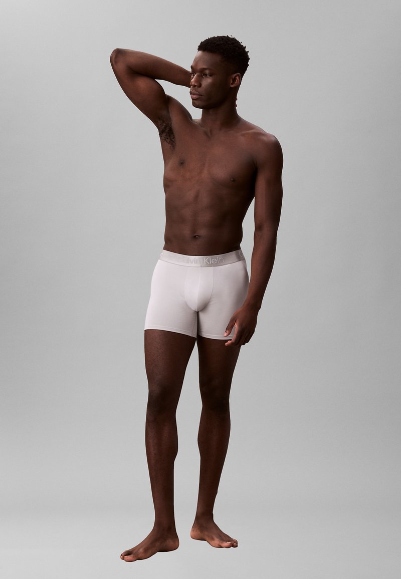 Boxers ajustados de algodón blanco con una suave cinturilla elástica que presenta el logo "Calvin Klein". El modelo está descalzo y de pie frente a un fondo gris.