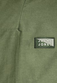 Tessuto verde oliva con una superficie liscia; include una toppa rettangolare nera con "JACK & JONES" impresso in bianco. Dettaglio di cucitura visibile.