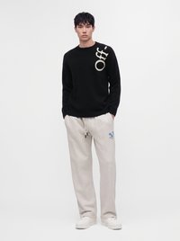 Giovane uomo che indossa un maglione nero con il testo "Off-" sulla spalla e pantaloni della tuta grigio chiaro con logo blu, in piedi con le mani in tasca.