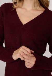 Pull en tricot bordeaux avec un col en V et des poignets côtelés. Texture douce, épaisseur moyenne, silhouette ajustée et légère variation de couleur.