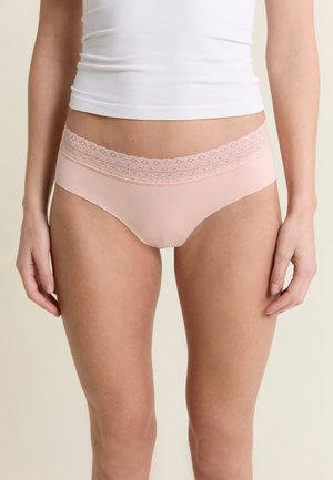 Mujer con bragas tipo hipster de color rosa claro con cintura de encaje y camiseta sin mangas blanca, de pie frente a un fondo neutro.