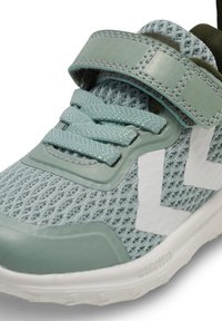 Scarpa sportiva azzurro chiaro con tomaia in mesh, suola in gomma e cinturino in Velcro. Presenta accenti bianchi a zig-zag sul lato e dettagli testurizzati.