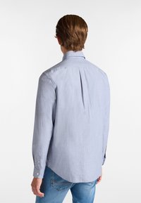 Camicia a maniche lunghe azzurro chiaro con placca, polsini a bottoni e orlo arrotondato. Il tessuto appare liscio con una sottile texture.