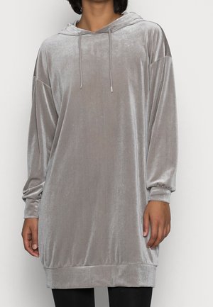 Osoba ubrana w luźną, długorękawową, szarą welurową sukienkę z kapturem i sznurkami, stojąca na tle jednolitego tła.