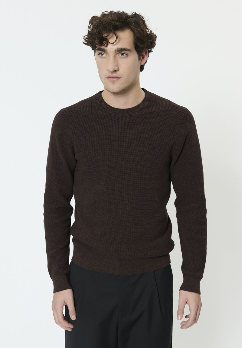 Pull marron en tricot avec un col rond et des manches longues. Texture côtelée aux poignets et à l'ourlet. Porté avec un pantalon foncé sur un fond uni.