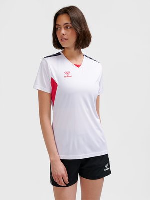 Kvinde iført en hvid sportstrøje med røde accenter og sorte shorts, kigger mod venstre mod en ensfarvet lys baggrund.