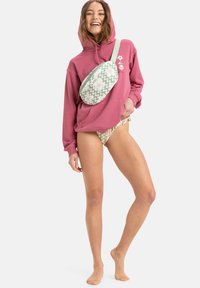 Różowa bluza, wzorzysty zielono-biały nerka, lekki bikini. Modelka stoi boso, zrelaksowana w pozie i z delikatnym uśmiechem.