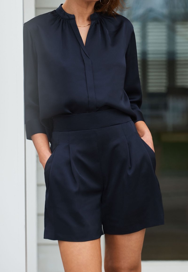 Navy blue satin blouse with a stand collar, pleated front, and three-quarter sleeves, paired with matching tailored shorts featuring side pockets.