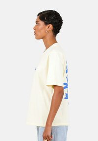 Cremefarvet bomulds t-shirt med korte ærmer, der har et blåt tropisk design på ryggen. Lige snit, afslappet pasform, minimal tekstur.