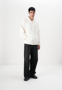 Hellbeige Kapuzenpullover mit einer Kängurutasche, Kordelzug und dezentem Branding, kombiniert mit dunkelblauen, weit geschnittenen Jeans und schwarzen Schuhen.