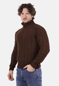 Maglione girocollo in maglia a treccia marrone con polsini e orlo a coste, caratterizzato da un motivo testurizzato e un design aderente. Indossato con jeans blu.