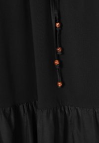 Tissu noir avec des détails froncés à la couture et un cordon noir suspendu avec quatre perles en bois marron régulièrement espacées.