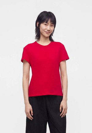 Personne aux cheveux noirs et raides sourit, portant un t-shirt rouge uni à manches courtes et un pantalon noir, sur un fond clair uni.