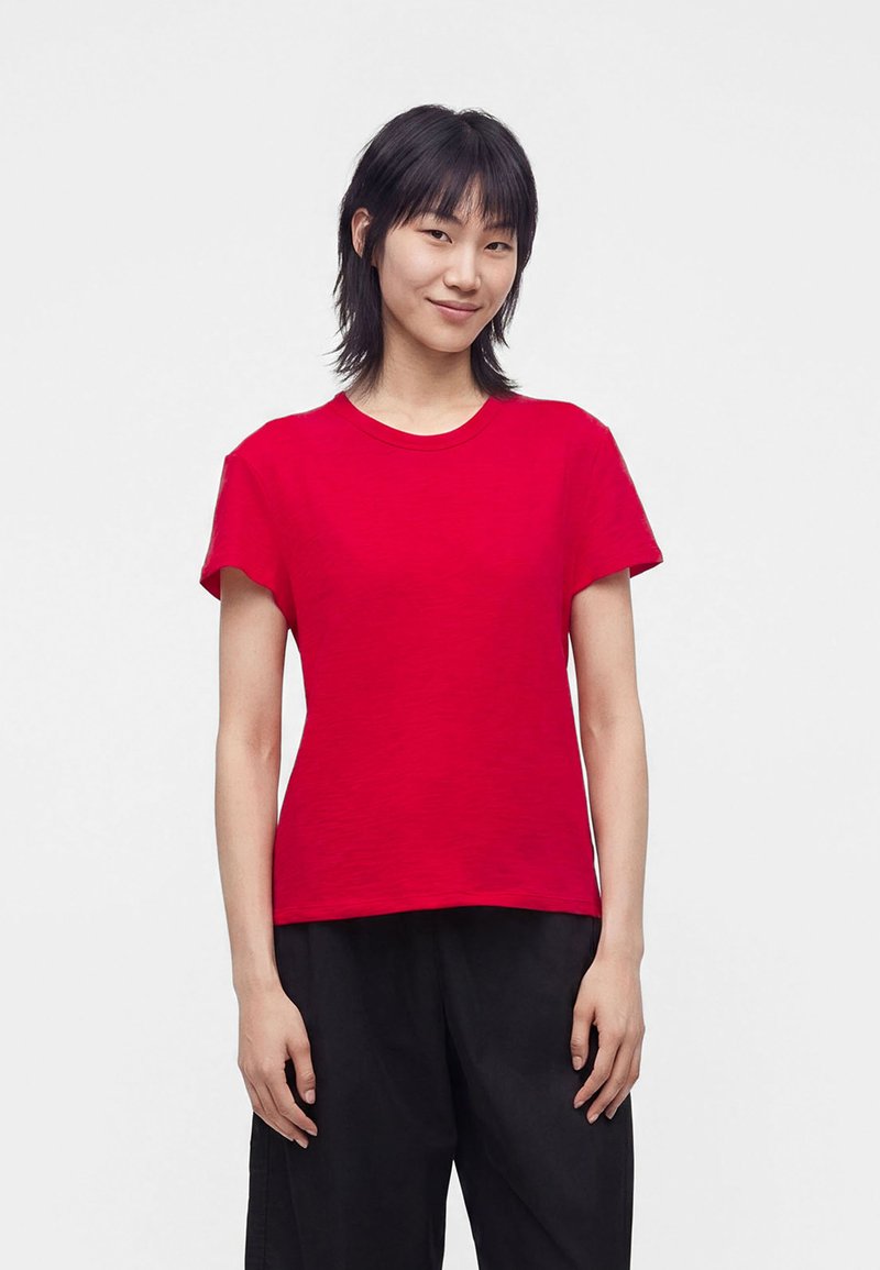 Personne aux cheveux noirs et raides sourit, portant un t-shirt rouge uni à manches courtes et un pantalon noir, sur un fond clair uni.