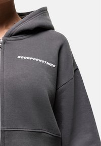 Grijze zip-up hoodie van zacht materiaal met een relaxte pasvorm, met het "GOODFORNOTHING" logo in het wit op de borst en een trekkoordkap.
